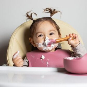 Small cute little toddler brunette caucasian girl with two tails tasting and enjoying by herself with a spoon the greek yogurt sitting in baby chair with messy face;